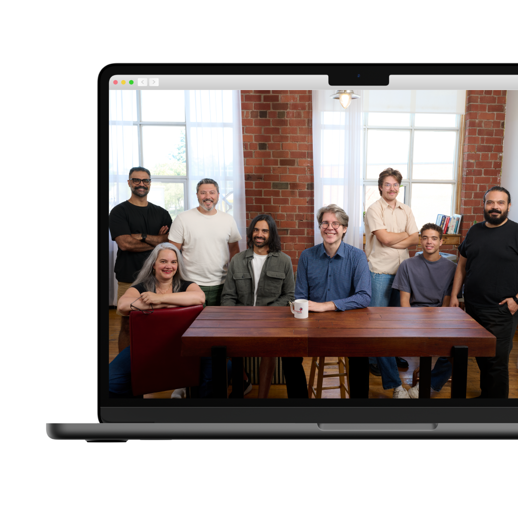 A group of eight people posing for a team photo in a bright office with brick walls and a large wooden table.