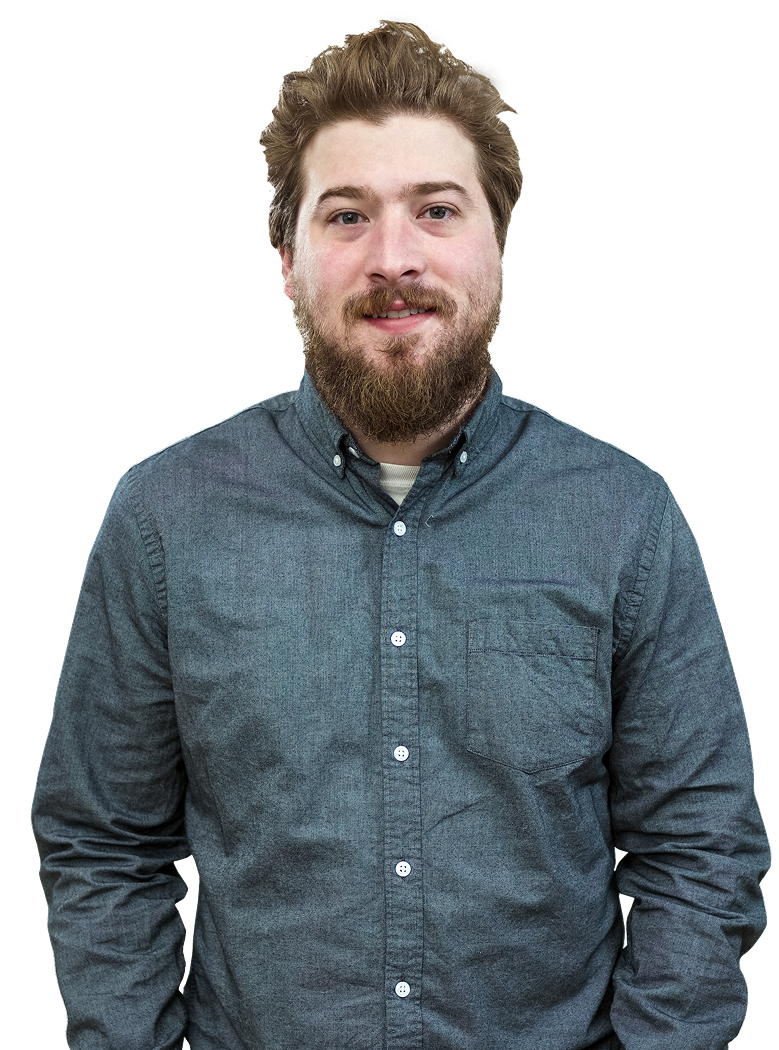 A man with a beard and brown hair wearing a grey button-down shirt. He is smiling and posing with hands in his pockets.
